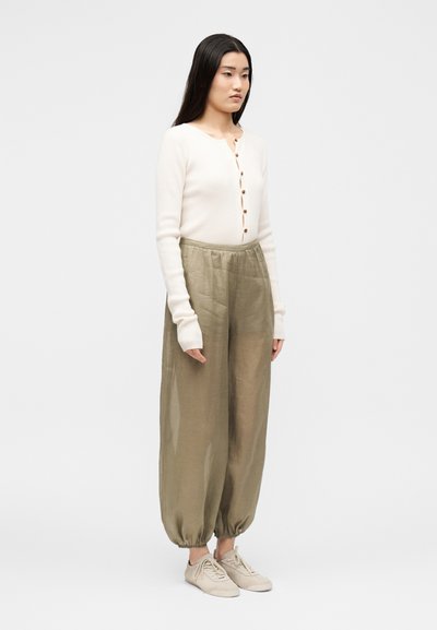 Femme debout portant un haut blanc à manches longues boutonné, un pantalon ample vert olive resserré aux chevilles et des baskets beige clair.