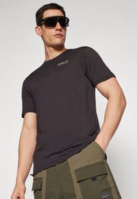 T-shirt a maniche corte grigio scuro con scollo rotondo, con un piccolo logo bianco sul petto. Abbinato a pantaloni cargo verde oliva.