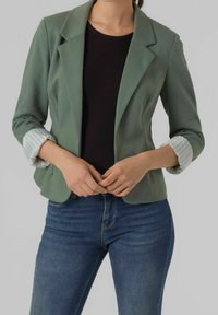 Veste verte au design structuré, avec des manches à rayures retroussées, portée sur un top noir et un jean bleu. Tissu lisse, coupe ajustée, longueur moyenne.