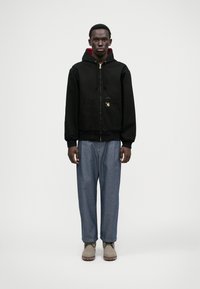 Moški v črni zadrgo zapiranju jopici z rdečim podstavkom, ohlapnih modrih kavbojkah in svetlo sivo obutvijo, stoji proti enobarvni beli ozadju.