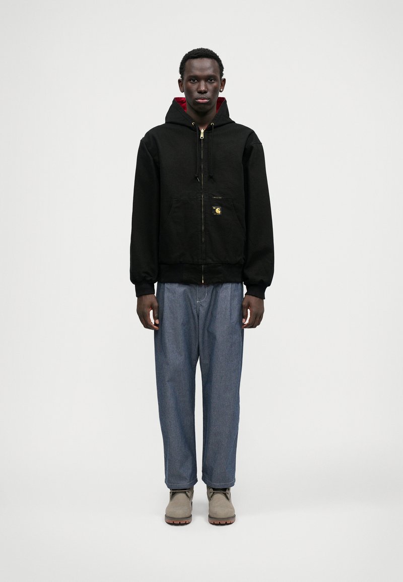 Moški v črni zadrgo zapiranju jopici z rdečim podstavkom, ohlapnih modrih kavbojkah in svetlo sivo obutvijo, stoji proti enobarvni beli ozadju.