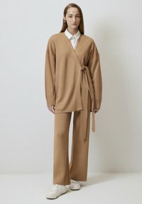 Beżowy sweter z owijanym paskiem, zestawiony z dopasowanymi spodniami o szerokich nogawkach i białymi sneakersami. Gładka faktura i luźny krój.