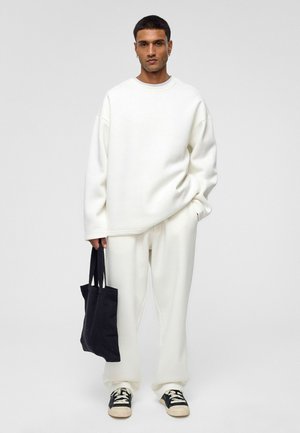 Witte oversized sweatshirt en bijpassende broek van zachte stof, gecombineerd met een zwarte tote bag en zwart-witte sneakers.