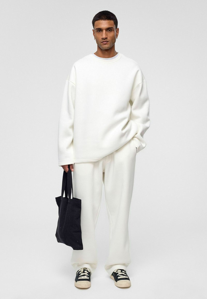 Witte oversized sweatshirt en bijpassende broek van zachte stof, gecombineerd met een zwarte tote bag en zwart-witte sneakers.