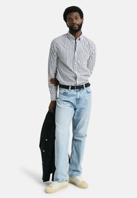 Chemise à rayures blanche et marine, jean bleu clair et baskets crème, tenant un blazer marine avec des boutons dorés.