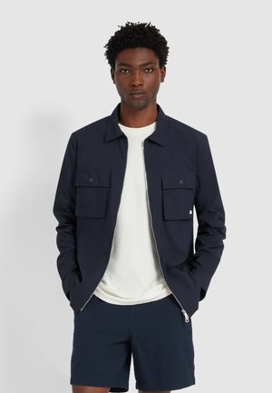 Marineblaue Jacke mit Reißverschluss, zwei Brusttaschen und Kragen. Über einem cremefarbenen Hemd getragen, kombiniert mit strukturierten marineblauen Shorts.