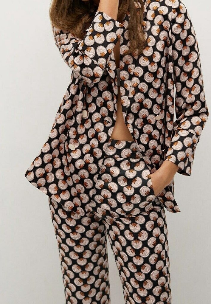 Femme portant un tailleur-pantalon assorti avec une base noire ornée d'un motif floral circulaire blanc, orange et marron, une main dans la poche et l'autre près du visage.