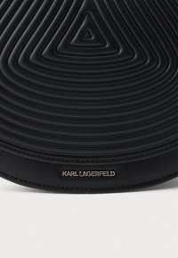 Μαύρη δερμάτινη τσάντα ώμου με υφή, προσφέρει σχέδιο με συντεταγμένους κυκλικούς κύκλους και χρυσό λογότυπο, υπογραμμίζοντας μια μοντέρνα και κομψή εμφάνιση.