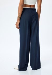 Marineblaue weit geschnittene Hose mit glatter Textur, ausgestattet mit einem verdeckten Bund und einer Gesäßtasche. Kombiniert mit weißen Sneakers.