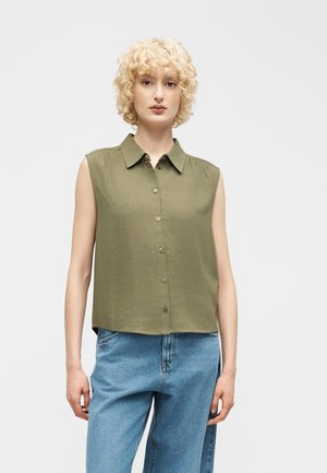 Žena s kučeravými blond vlasmi, oblečená v olivovo zelenej bezrukávovej košeli na gombíky a svetlomodrých džínsoch, stojaca pred jednoduchým svetlým pozadím.