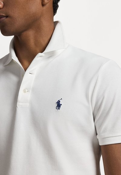 Biely polo tričko vyrobené z textúrovanej látky, s námorníckym modrým vyšitým logom na ľavej hrudi a klasickým dizajnom goliera.