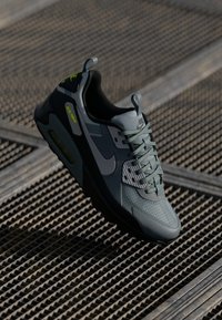 Nike Air Max shoe in grey with mesh panels, textured surfaces, green accents, and a cushioned sole. Features prominent logo and lacing.