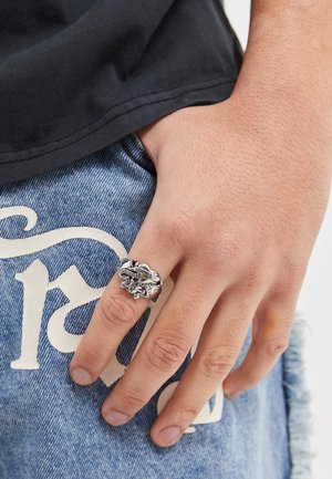 Silver ring with intricate floral and snake design, featuring a polished finish. Worn on the index finger, against blue denim shorts.