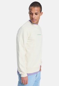 Sweat-shirt crème clair avec un col rond, des manches longues et un logo vert. Superposé sur une chemise violette, avec une coupe décontractée et une texture douce.