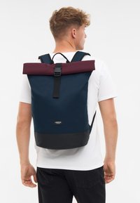 Sötétkék, felgöngyölhető hátizsák, bordó kiegészítővel és fekete alappal. Tartós anyagból készült, állítható pántokkal és elülső csatbezárással.