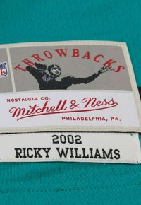 Étiquette en tissu vert avec surface texturée ; présente le mot "Throwbacks" en rouge et "Mitchell & Ness" en cursive, ainsi que "2002" et "Ricky Williams" en noir.
