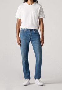 Vit bomullströja med bröstficka, kombinerad med medelblå jeans. Modellen bär vita sneakers. Enkel, avslappnad outfit.