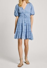 Jasnoniebieska sukienka z dekoltem w kształcie litery V, bufkami na rękawach i talią z troczkiem. Posiada wzory z ażurowych otworów i falowany dół. Zestawiona ze złotymi sandałami.