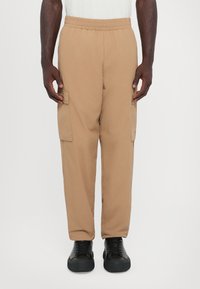 Pantalons cargo beiges avec ceinture élastique, dotés de poches latérales et d'une coupe décontractée, associés à des chaussures noires à lacets.