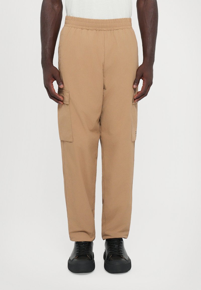 Pantalons cargo beiges avec ceinture élastique, dotés de poches latérales et d'une coupe décontractée, associés à des chaussures noires à lacets.