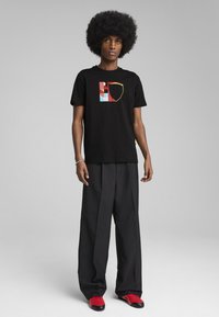 Sort bomuld t-shirt med et grafisk design i rød, blå og gul. Båret med løse sorte bukser og røde sko.