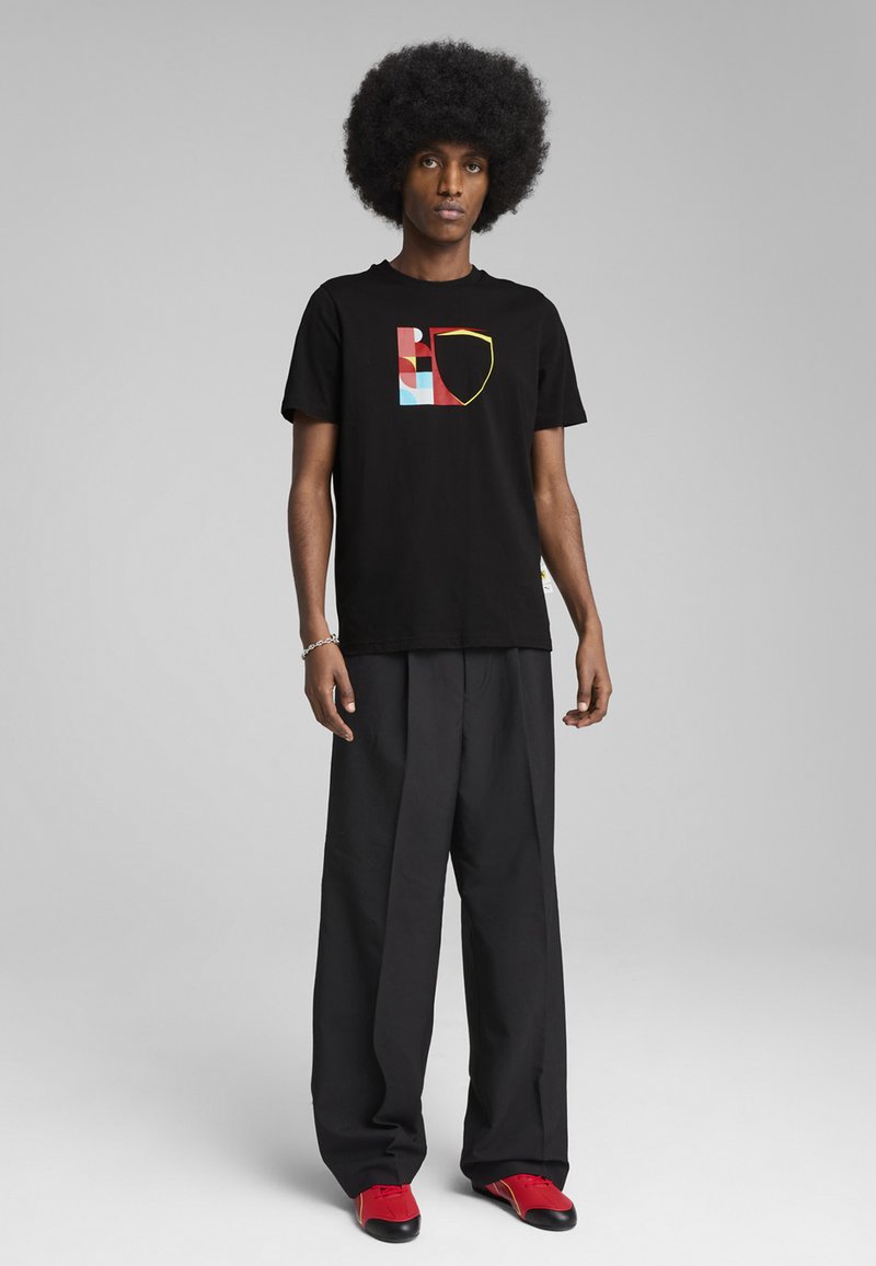 Sort bomuld t-shirt med et grafisk design i rød, blå og gul. Båret med løse sorte bukser og røde sko.