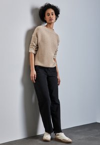 Beige und cremefarben gestreifter Pullover mit Dreiviertelärmeln, schwarze gerade Jeans und beigem Sneakers vor einer hellblauen Wand.