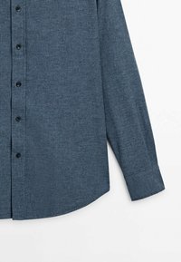 Chemise bleue à manches longues avec petit motif à carreaux, montrant les boutons devant et le poignet sur fond blanc.