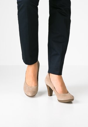 Kvinde iført marineblå bukser og beige ruskind højhælede pumps står på en hvid gulv med den ene fod let fremme.