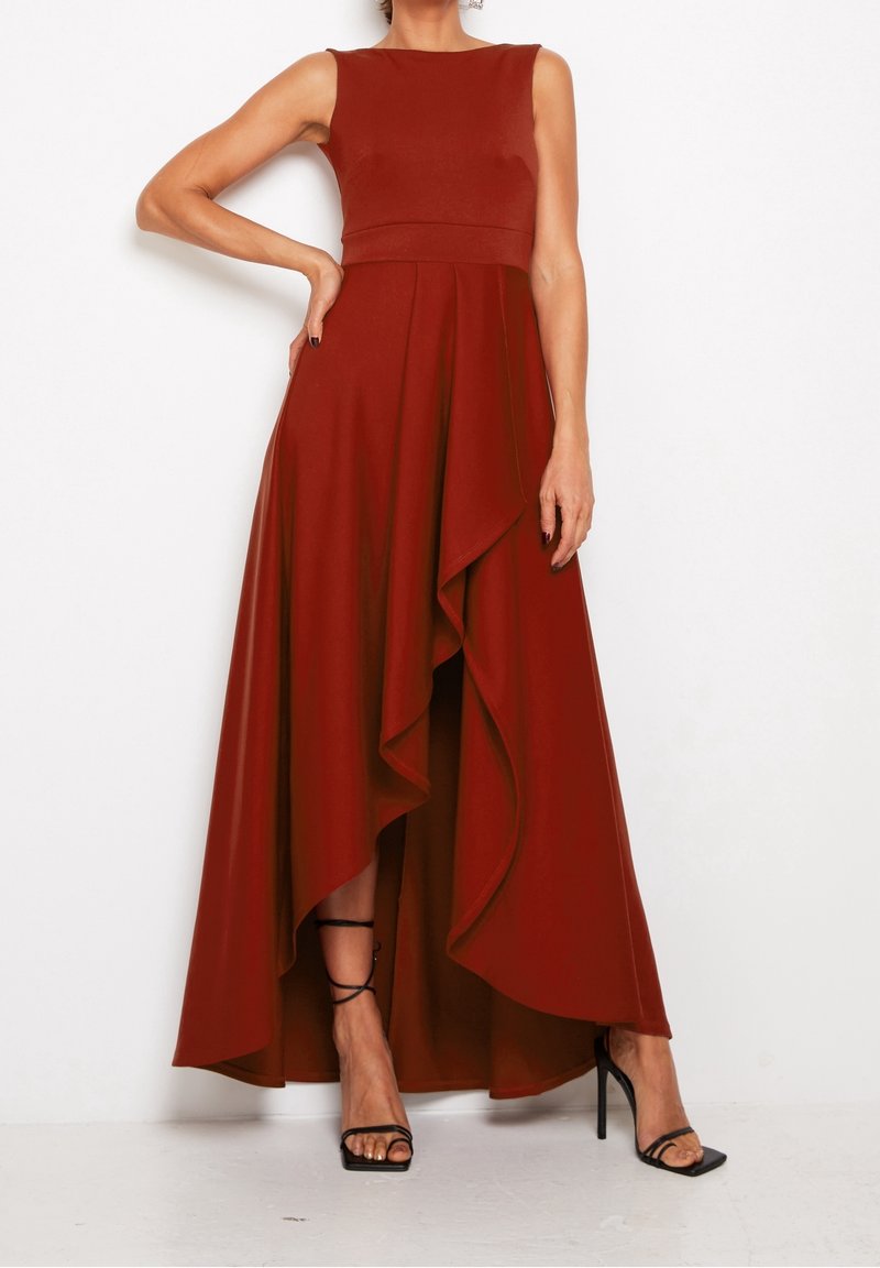 Femme portant une robe sans manches rouge rouille à jupe asymétrique avec un détail volanté et des sandales noires à talons hauts et lanières, debout sur fond blanc.