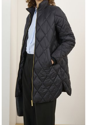 Frau trägt eine schwarze gesteppte Jacke mit goldfarbenem Reißverschluss über einem hellblau gestreiften Hemd und schwarzen Hosen, mit der Hand in der Jackentasche.