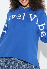 Felpa con cappuccio blu con maniche lunghe e scritta bianca "Travel Vibe." Tessuto morbido, vestibilità comoda e cappuccio con coulisse. Indossata sopra pantaloni con motivo.