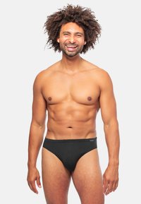 Zwarte mesh zwemslips met een lage taille, een gemarmerd patroon en een comfortabele tailleband. Het model staat rechtop tegen een eenvoudige achtergrond.