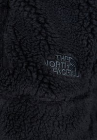 Pull noir en polaire avec une surface texturée. Comprend une poche zippée et un logo brodé gris foncé sur la poitrine.