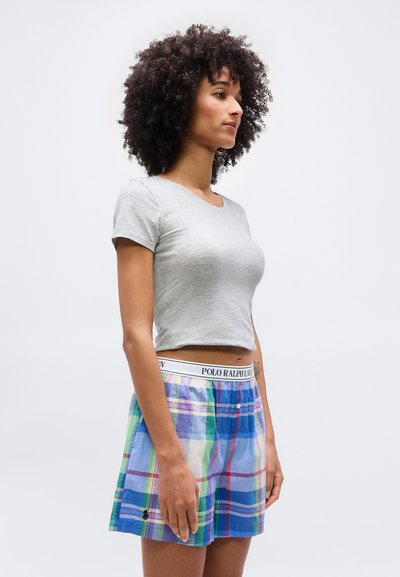 Γυναίκα με σγουρά μαλλιά, φορώντας ανοιχτό γκρι cropped t-shirt και πολύχρωμα καρό σορτς με ζώνη Polo Ralph Lauren, όρθια σε προφίλ.
