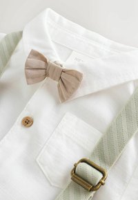 Camicia bianca con cravatta a farfalla beige, bottone di legno e bretelle a righe verdi fissate con fermagli di ottone. Dettagli di texture semplici.