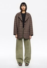 Blazer de cuadros marrón de corte oversize, top de punto oscuro, pantalones de pierna ancha en color oliva y sandalias negras con detalles florales. Diseñado con un ajuste relajado.