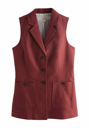 Donna con blazer bordeaux senza maniche e pantaloncini tiene una borsa floreale, in piedi vicino a un muro bianco con oceano e vegetazione sullo sfondo.