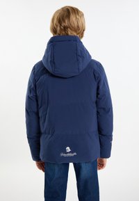 Temno moder puffer plašč z kapuco, s hladko, prešito teksturo in logotipom na robu. V kombinaciji z modrimi kavbojkami.