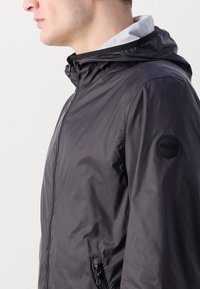 Jachetă ușoară neagră cu glugă, din material nylon, cu fermoar frontal, buzunare laterale și un patch rotund cu logo pe mânecă stângă.