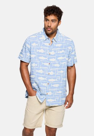 Hombre vestido con una camisa de manga corta color azul claro con patrones de peces blancos y pantalones cortos beige remangados, de pie con una mano en el bolsillo.