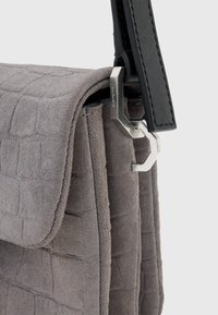 Grå ruskinds håndtaske med et struktureret krokodillemønster, udstyret med en sort læderrem og sølvmetal hardware. Lukket klapdesign.