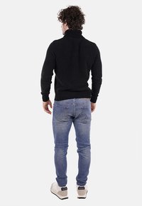 Maglione nero a coste con colletto alto, indossato con jeans azzurri chiari e sneakers beige. Il maglione presenta un design aderente e un tessuto testurizzato.