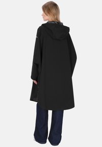 Poncho de ploaie negru cu glugă, design lung și lejer, prevăzut cu închideri cu capse pe părțile laterale și o textură fină. Purtați peste blugi bleumarin.