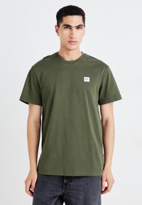 Olijfgroene katoenen t-shirt met een ronde hals, korte mouwen en een klein rechthoekig label op de linker borst. Eenvoudig ontwerp.