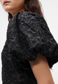 Črna bluzica z teksturo, ki ima napihnjene rokave, subtilen reliefni vzorec in strukturiran kroj, kar poudarja njeno edinstveno zasnovo tkanine.