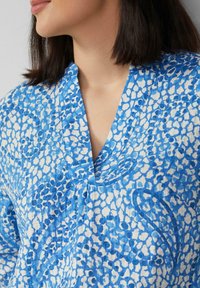 Blauw-witte blouse met patroon, voorzien van een V-hals ontwerp en een gestructureerde stof. Het patroon bevat verschillende abstracte vormen en accenten.