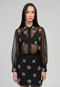 Blouse noire transparente à manches longues, ornée de motifs floraux brodés en blanc et en argent, avec un devant à boutons. Associée à une jupe assortie.