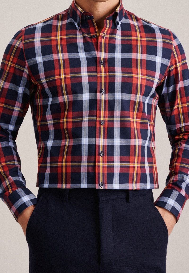 Mężczyzna w czerwonej, granatowej, żółtej i białej koszuli w kratkę z guzikami, w granatowych spodniach, z rękami w kieszeniach, widoczny tors, na jednolitym tle.