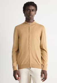 Man in een beige ritsvest en witte broek, staande tegen een plain witte achtergrond, rechtop met een neutrale uitdrukking.